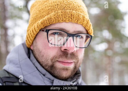 Portrait von ein ernster Mann mit Bart und Brille mit einer unerschütterlichen Blick gegen einen unscharfen Hintergrund schneebedeckter Wald an einem frostigen Wintertag Stockfoto