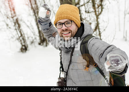 Winter Spaß. Freudige aussehenden bärtigen Mann in Gläsern mit einer Kamera und einem Rucksack bereitet einen Schneeball zu werfen, unter Schneeverwehungen und Wald. Stockfoto