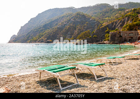 Monterosso al Mare, Italien - September 02, 2019: Grüne Liegestühle auf Kieselstrand mit grünen Bergen im Hintergrund in Monterosso, Cinque Stockfoto