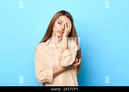 Schöne erschöpft Frau schläfrig fühlt, deckt das Gesicht mit Palm, seufzt von Müdigkeit, trägt Mode Shirt, auf blauem Hintergrund. Negative feelin Stockfoto