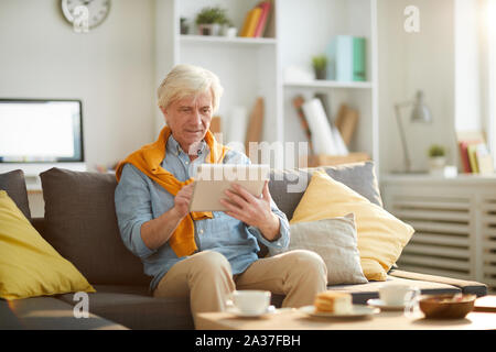 Portrait des modernen älterer Mann mit digitalen Tablet und entspannen Sie sich auf bequemen Couch Platz zu Hause, kopieren Stockfoto