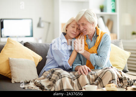 Portrait der liebevollen Umarmung senior Paar zärtlich auf bequemen Couch zu Hause sitzen, mit warmen Decke zugedeckt, kopieren Raum Stockfoto