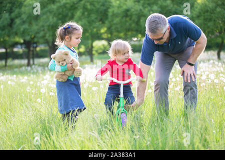 Großvater lehrt Enkelkinder eine Radtour. Gerne ältere Großvater mit zwei Kindern liegen auf Gras im Park Stockfoto