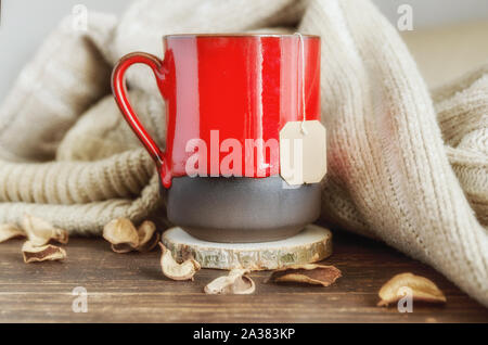 Rot Tasse Kräutertee, beige gemütlich gestrickten Pullover und trockene Blätter auf dem hölzernen Hintergrund. Köstlich kaltem Wetter Trinken für den Herbst. Aufenthalt im Ho Stockfoto