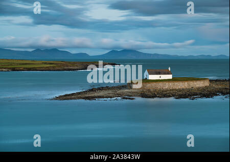 Einem stimmungsvollen Sonnenuntergang an der historischen St Cwyfan Kirche, Anglesey, befindet sich auf der kleinen Insel Gezeiten an der Küste von Cribinau - "Kirche am Meer" Stockfoto