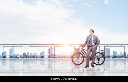 Junger Mann mit Anzug stehen auf Balkon mit dem Fahrrad. Geschäftsmann mit dem Fahrrad auf dem Hintergrund des blauen Himmels über Megalopolis. Männliche Radfahrer halten Stockfoto