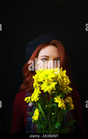 Französische rothaarige Frau in karierten Kleid, Burgund Pullover und schwarzen Barett hält und Schnüffelt gelbe Chrysanthemen Stockfoto