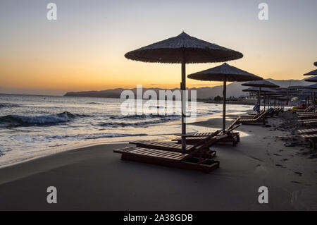 Sonnenliegen und Sonnenschirme bei Sonnenaufgang am Strand von Stalis im Norden von Kreta Stockfoto