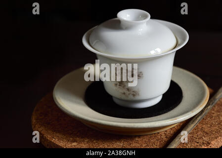 Nahaufnahme eines isolierten Chinesische Weisse Teekanne mit Tee und Deckel, die steht auf einer Platte und auf einem Kork Untersetzer mit einem gebrauchten Essstäbchen nächsten t Stockfoto