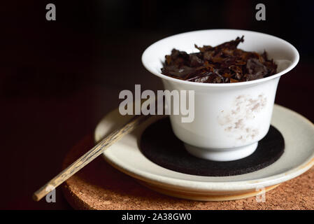 Nahaufnahme eines typischen chinesischen Porzellan Teekanne in Weiß mit gebrühter schwarzer Tee Blätter, es auf eine Untertasse und ein Kork Platte mit einer alten hölzernen Stock gege Stockfoto