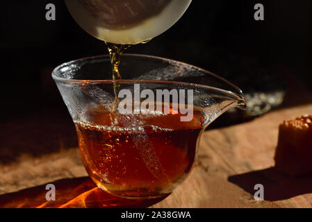 Nahaufnahme einer Teekanne aus Glas mit schwarzen frischen Tee, in denen Kaffee aus einem anderen Kaffee gegen einen dunklen Hintergrund auf einen Tisch geschüttet wird an einem Stockfoto