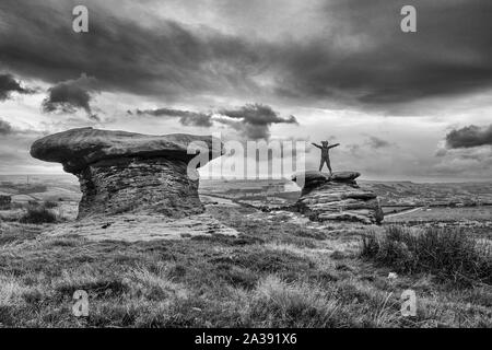 Person, die auf der Oberseite des Doppler Steine mit ausgestreckten Armen, Ilkley Moor, UK ihre ungewöhnliche Form, indem Sie die Oberseite der Festplatte grits verursacht wird Stockfoto