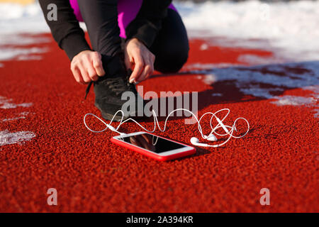 Mädchen Athlet in schwarzen Sneakers, geduckt auf der roten Spur ausgeführt werden. In der Nähe ist ein Telefon mit drahtgebundenen Kopfhörern. Kalten verschneiten Wetter. Stockfoto