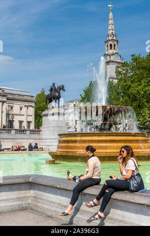 Zwei junge Mädchen genießen den Sonnenschein sitzen auf dem Rand eines Brunnen am Trafalgar Square, Central London. Stockfoto