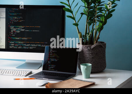 Zwei schwarze Bildschirme, Laptop und Monitor, mit Programmcode Linien auf dem Schreibtisch gefüllt. Stockfoto