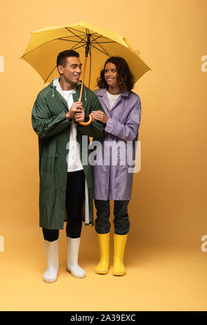 Lächelnd interracial Paare in Regenmäntel und Gummistiefel holding Umbrella auf gelbem Hintergrund Stockfoto