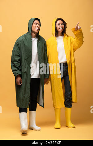 Happy interracial Paare in Regenmäntel und Gummistiefel zeigt mit dem Finger auf gelbem Hintergrund Stockfoto