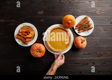 Teilweise mit Blick auf die männliche Hand in der Nähe der Platte mit Pumpkin Pie, gebackene ganze Karotte, in Scheiben geschnitten und ganze Kürbisse auf dunklen Holztisch Stockfoto