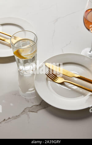 Glas mit Zitrone Wasser in der Nähe von weißen Platten mit goldenen Gabeln und Messer auf Marmor tisch Stockfoto