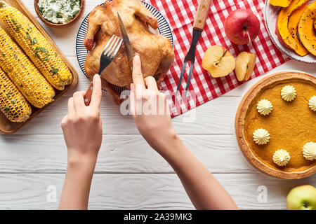 7/8-Ansicht von Frau schneiden gebratener Truthahn in der Nähe von Pumpkin Pie und gegrilltem Gemüse serviert auf weiße Holztisch mit Rot karierte Servietten Stockfoto