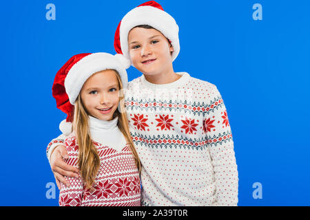 Zwei lächelnde Kinder in Pullover und Nikolausmützen isoliert, die auf Blau Stockfoto