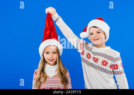 Zwei lächelnde Kinder in Pullover und Nikolausmützen isoliert auf Blau Stockfoto