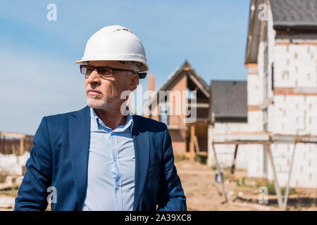 Reifen und bärtigen Geschäftsmann in Anzug und Helm in der Nähe von Gebäuden Stockfoto