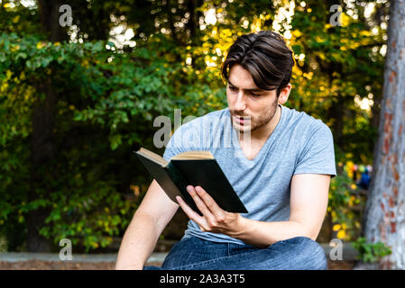 Junger Mann liest eine self-centered Roman in einem Park bei Sonnenuntergang sitzen, genießen der antiken Literatur. Stockfoto