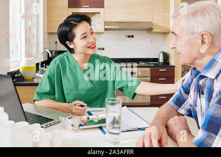 Freundliche hübsche Krankenschwester am Küchentisch der älteren Patienten sitzen und erklären ihm Art und Weise der Behandlung Stockfoto