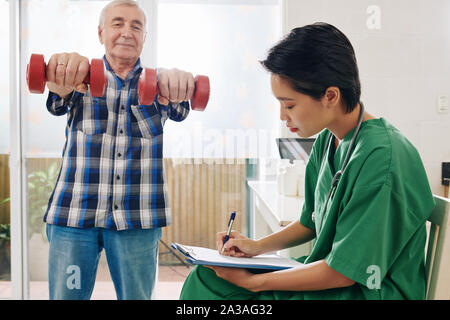 Lächelnd älterer Mann Übungen mit Hanteln, wenn Krankenschwester Notizen in medical card Stockfoto