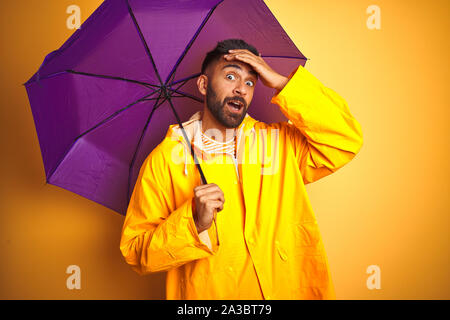 Junge indische Mann mit Regenmantel und Regenschirm über isolierte Gelb Lila Hintergrund mit Hand betonte am Kopf, mit Scham und Überraschung Gesicht schockiert Stockfoto