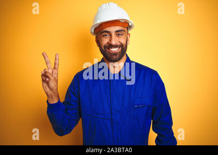 Hübscher indischer Arbeitnehmer Mann einheitliche und Helm tragen über isolierte gelben Hintergrund lächelnd mit glücklichen Gesicht in die Kamera winking tun Sieg Zeichen Stockfoto