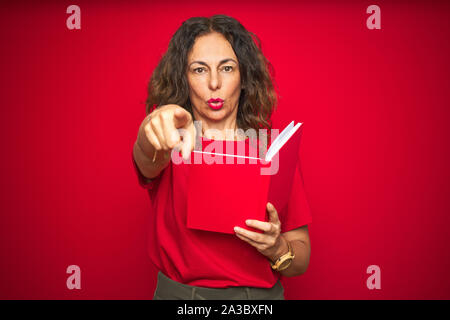 Mittleren Alters ältere Frau mit einem Buch über den roten Hintergrund isoliert zeigte mit dem Finger an die Kamera und an Sie, Hand unterzeichnen, positiv und zuversichtlich g Stockfoto