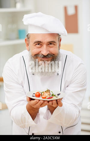 Portrait von bärtigen Senior Chef präsentiert schöne italienische Gericht und glücklich lächelnd in die Kamera während im Restaurant Küche posing Stockfoto