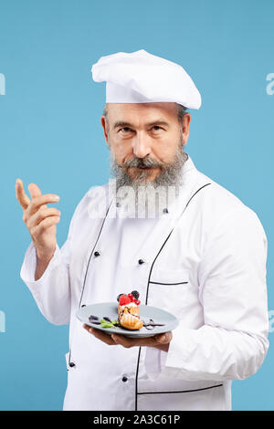 Taille bis Porträt des charismatischen bärtigen Koch präsentiert schöne Dessert beim Stehen vor blauem Hintergrund Stockfoto