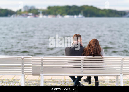 Junges Paar entspannende auf einer Promenade am Wasser sitzen zusammen auf einem weissen Sitzbank mit Blick auf kabbelwasser an einem kalten und windigen Herbst Tag vom gesehen Stockfoto