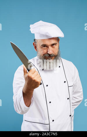 Taille bis Porträt des charismatischen bärtigen Koch Holding scharfen Messer und Kamera beim Stehen vor blauem Hintergrund Stockfoto
