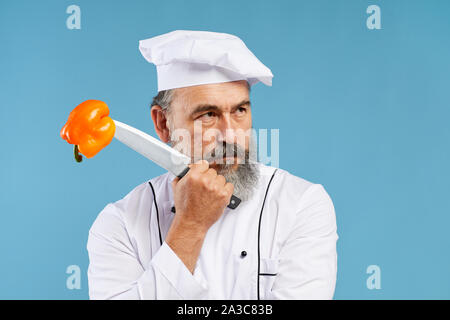 Portrait von nachdenklich bärtigen Koch Holding scharfe Messer mit Paprika durchbohrt auf Ihre IT, während gegen den blauen Hintergrund im Studio Posing, kopieren Raum Stockfoto