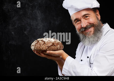Porträt des charismatischen bärtigen Bäcker mit frischem Brot Brot während auf schwarzem Hintergrund posiert und lächelnd an Kamera, kopieren Raum Stockfoto