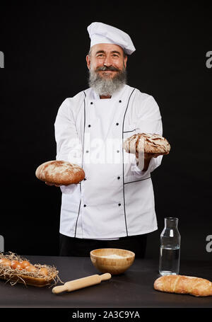 Portrait von bärtigen Bäcker Holding frisches Brot glücklich lächelnd an der Kamera, während auf schwarzem Hintergrund posiert Stockfoto