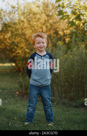 Portrait der kaukasischen Rothaarige niedlich aussehenden Vorschule Junge. Adorable Kind stehen draußen auf Herbst Tag. Glücklich lächelnde Kid outdoor. Authentische Stockfoto