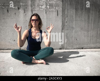 Mädchen in der Sonnenbrille ist das Meditieren über eine bituminöse Fußboden gegen eine graue Wand. Lifestyle, Identität mit Gott, das Bild einer urbanen Stil. Stockfoto