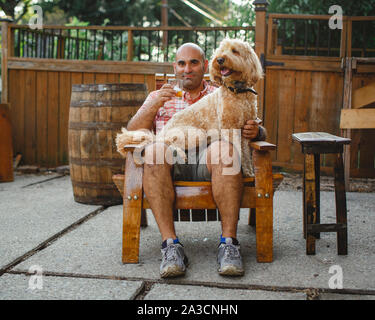 Ein glücklicher Mann sitzt mit Glas Bourbon und Hund auf dem Schoß auf hölzernen Stuhl Stockfoto