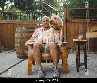 Ein Mann sitzt auf einer handgefertigten Holzstuhl mit einem glücklichen Hund auf seinen Schoß. Stockfoto