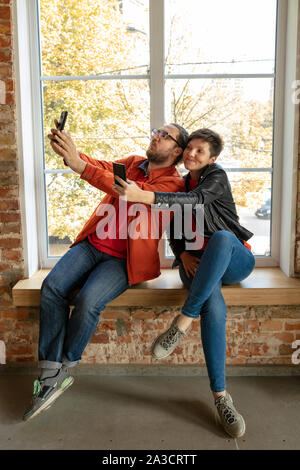 Happy kaukasischen Jugendlichen, Paar hinter dem Backstein Fenster. Gemeinsame Nutzung einer News, Fotos oder Videos von Smartphones, Laptops oder Tabletten, spielen und Spaß haben. Soziale Medien, moderne Technologien. Stockfoto