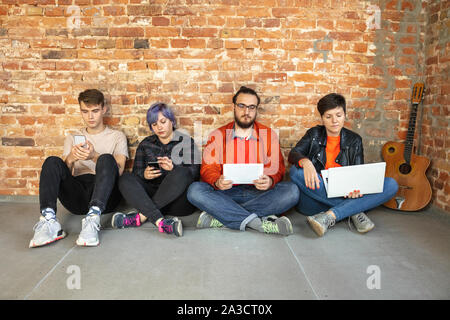 Gruppe von Happy kaukasischen jungen Menschen hinter der Mauer sitzen. Gemeinsame Nutzung einer News, Fotos oder Videos von Smartphones oder Tabletten, spielen und Spaß haben. Soziale Medien, moderne Technologien. Stockfoto