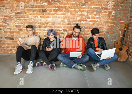 Gruppe von Happy kaukasischen jungen Menschen hinter der Mauer sitzen. Gemeinsame Nutzung einer News, Fotos oder Videos von Smartphones oder Tabletten, spielen und Spaß haben. Soziale Medien, moderne Technologien. Stockfoto