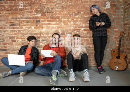 Gruppe von Happy kaukasischen jungen Menschen hinter der Mauer. Gemeinsame Nutzung einer News, Fotos oder Videos von Smartphones, Laptops oder Tabletten, spielen und Spaß haben. Soziale Medien, moderne Technologien. Stockfoto