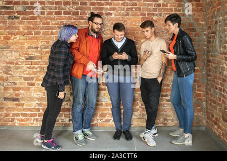 Gruppe von Happy kaukasischen jungen Menschen hinter der Mauer stehen. Gemeinsame Nutzung einer News, Fotos oder Videos von Smartphones oder Tabletten, spielen und Spaß haben. Soziale Medien, moderne Technologien. Stockfoto