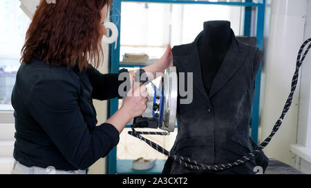 Nähen Produkte in Tailoring business. Nahtlose Zusammenarbeit mit dem Bügeln Weste auf Mannequin mit Dampf in der Schneiderei. Fashion Designer bei der Arbeit parow Kleidung mit Bügeleisen, close-up. Bekleidung Produktion. Stockfoto